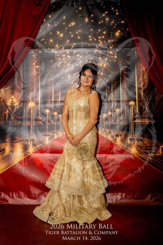 A person in a gold, floor-length gown stands before a red carpet backdrop with fairy lights for the 2026 Military Ball.