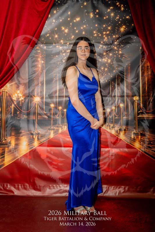A person in a blue satin gown stands on a red carpet in front of a backdrop for the 2026 Tiger Battalion Military Ball.