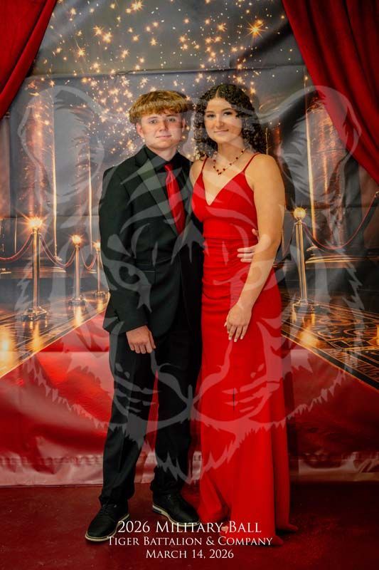 A couple in formal attire stands together for a 2026 military ball photo against a red carpet and curtain backdrop.