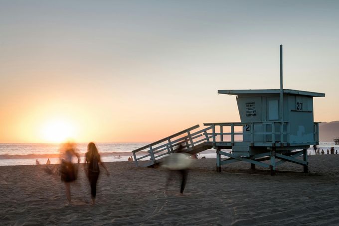 Beach scene at sunset; lifeguard stand, surfers with boards, blurred figures on sand.