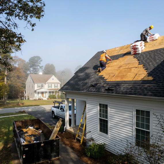 Roofers repairing storm-damaged shingles on a white house, with a dumpster and ladder below