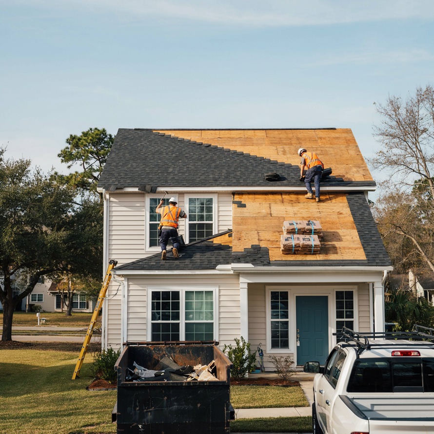 Workers repairing a house roof and siding beside a white pickup truck and dumpster