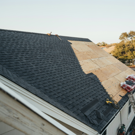 Roof under construction with dark shingles, exposed plywood sections, and roofing materials on the right side