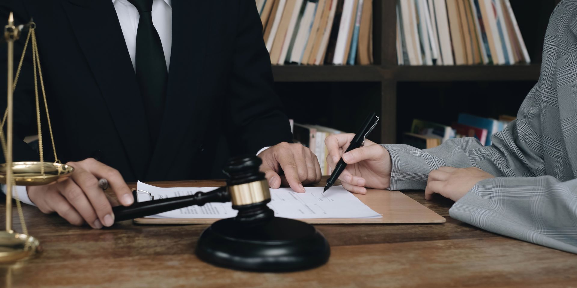 A person in a suit sits with another person, signing a document. A gavel and scale are on the table.