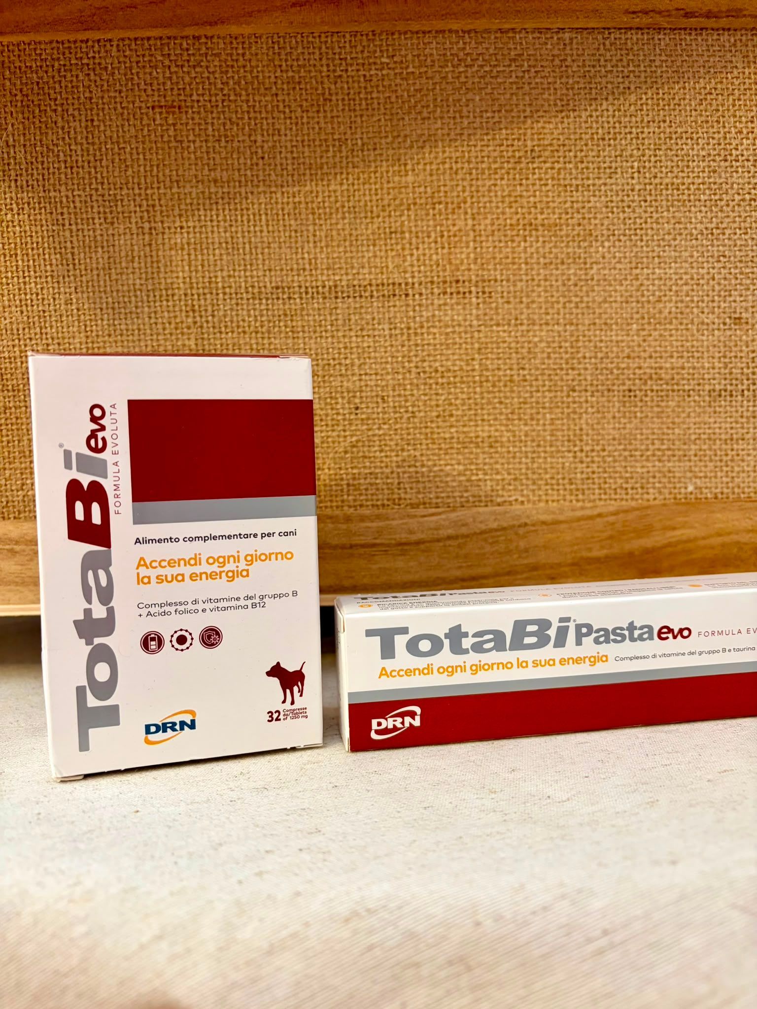 Scatole per medicinali TotaBio su una superficie beige intrecciata, con confezione rossa e bianca.