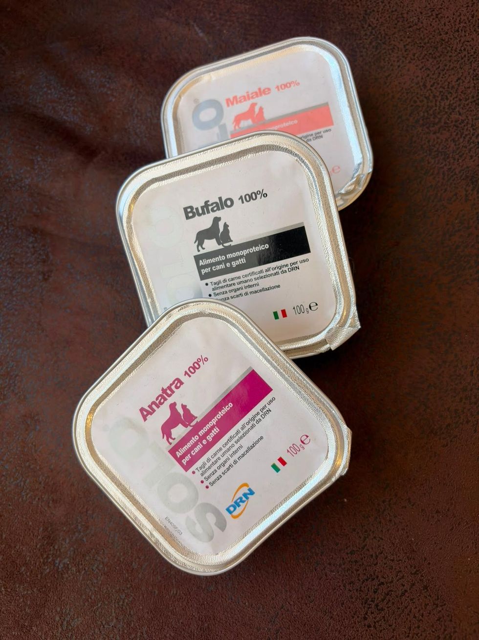 Tre contenitori quadrati per cosmetici impilati su una superficie marrone, con etichette bianche, nere e rosa.