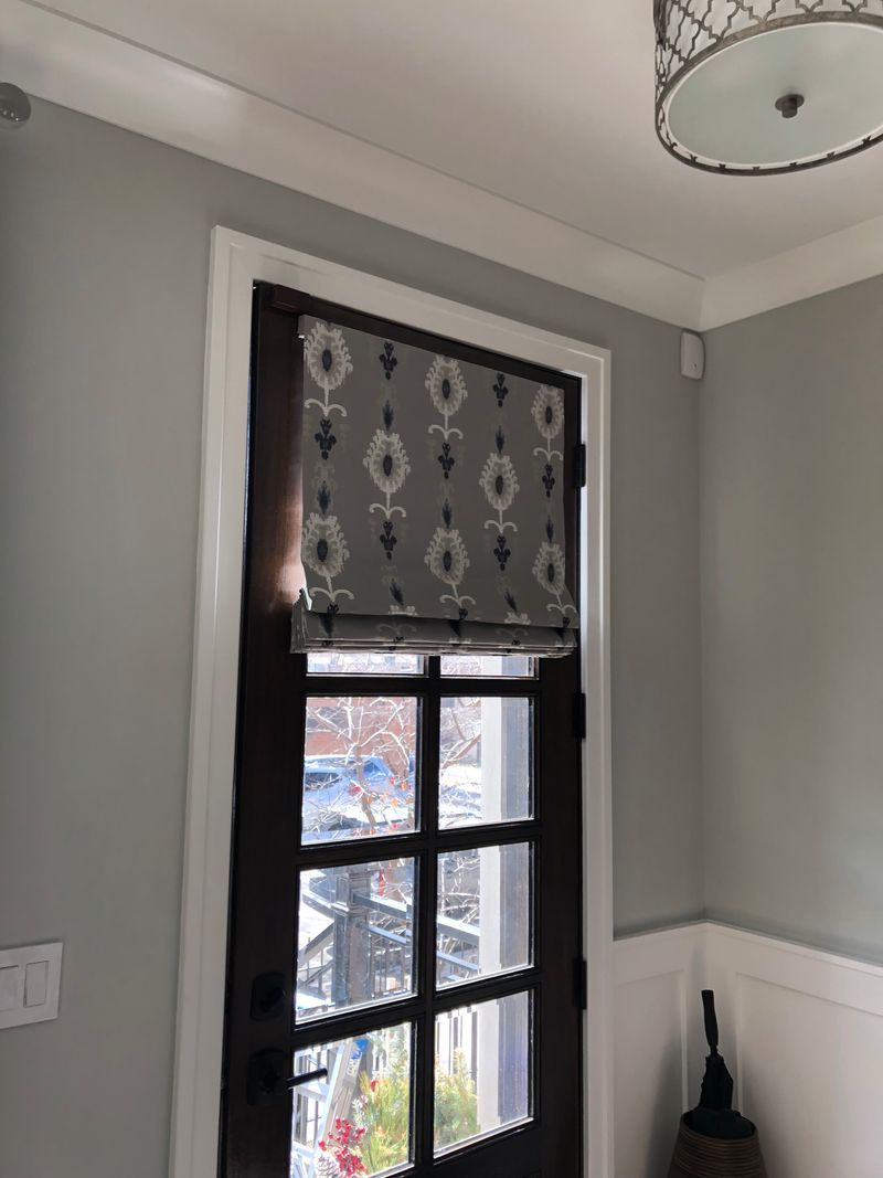 Black-framed window with patterned gray Roman shade in a light gray room, with white wainscoting and a ceiling light.