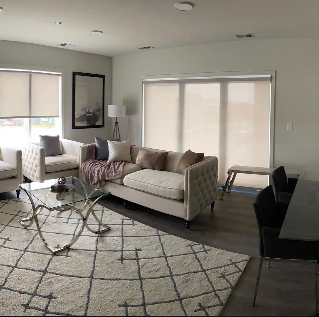 Bright living room with beige sofa, patterned rug, white chairs, and large windows with blinds