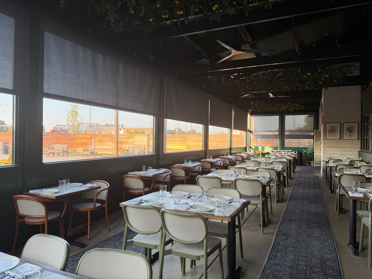 Empty restaurant dining area with set tables, white chairs, and large windows overlooking an outdoor view.
