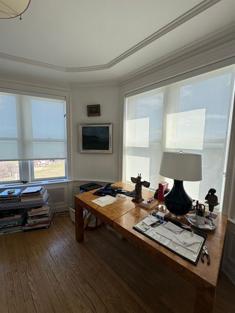 Bright corner office with wooden desk, large windows, lamp, and wall art overlooking a city view