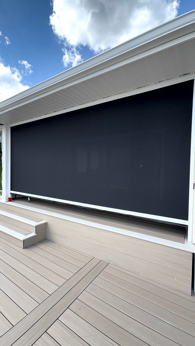 Outdoor deck with a large dark gray retractable screen under a white roof, beside blue sky and clouds