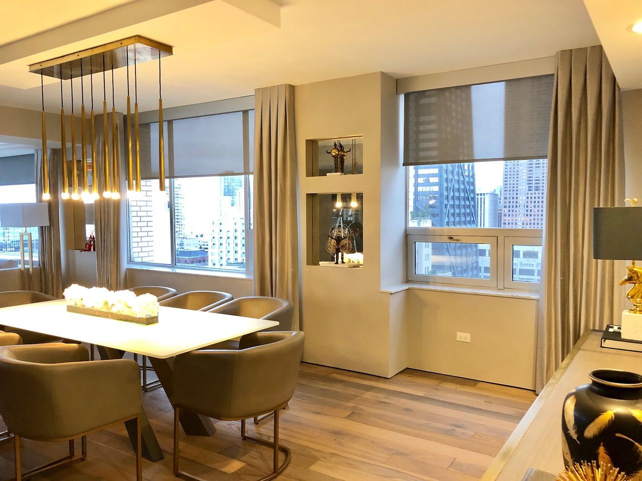 Modern dining room with large windows, beige chairs, a lit table, and city buildings outside