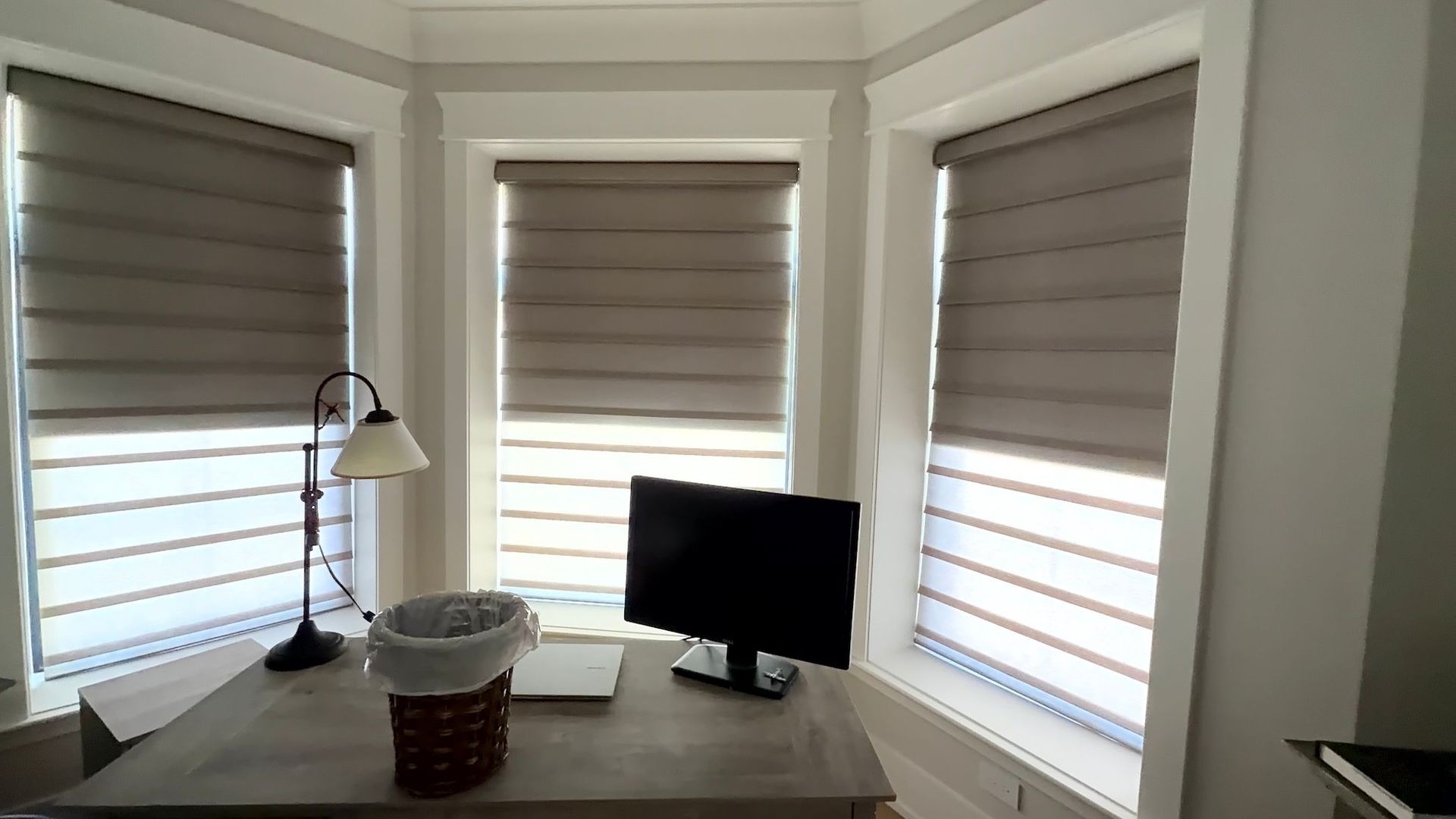 Home office desk by bay windows with beige blinds, lamp, monitor, and a basket on the table.