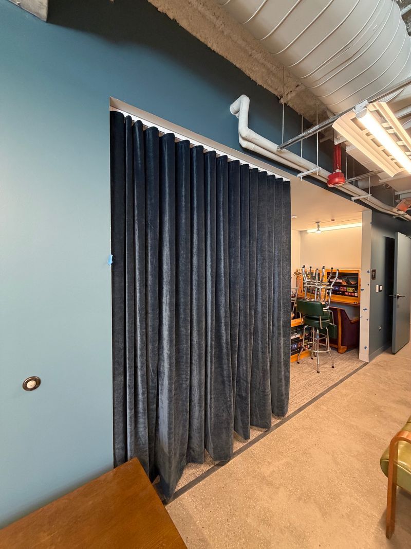 Office hallway with dark blue curtains, exposed ceiling ducts, and a glimpse into a plant-filled workspace