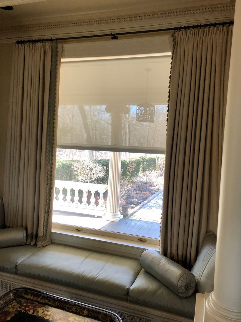 Living room window with beige curtains, cushioned bench, and a view of a sunny balcony outside