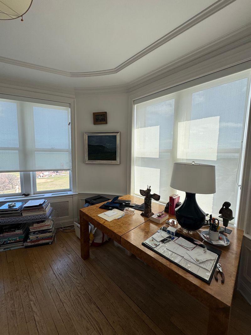 Bright home office with wooden desk, lamp, books, and large sunlit windows overlooking the city.