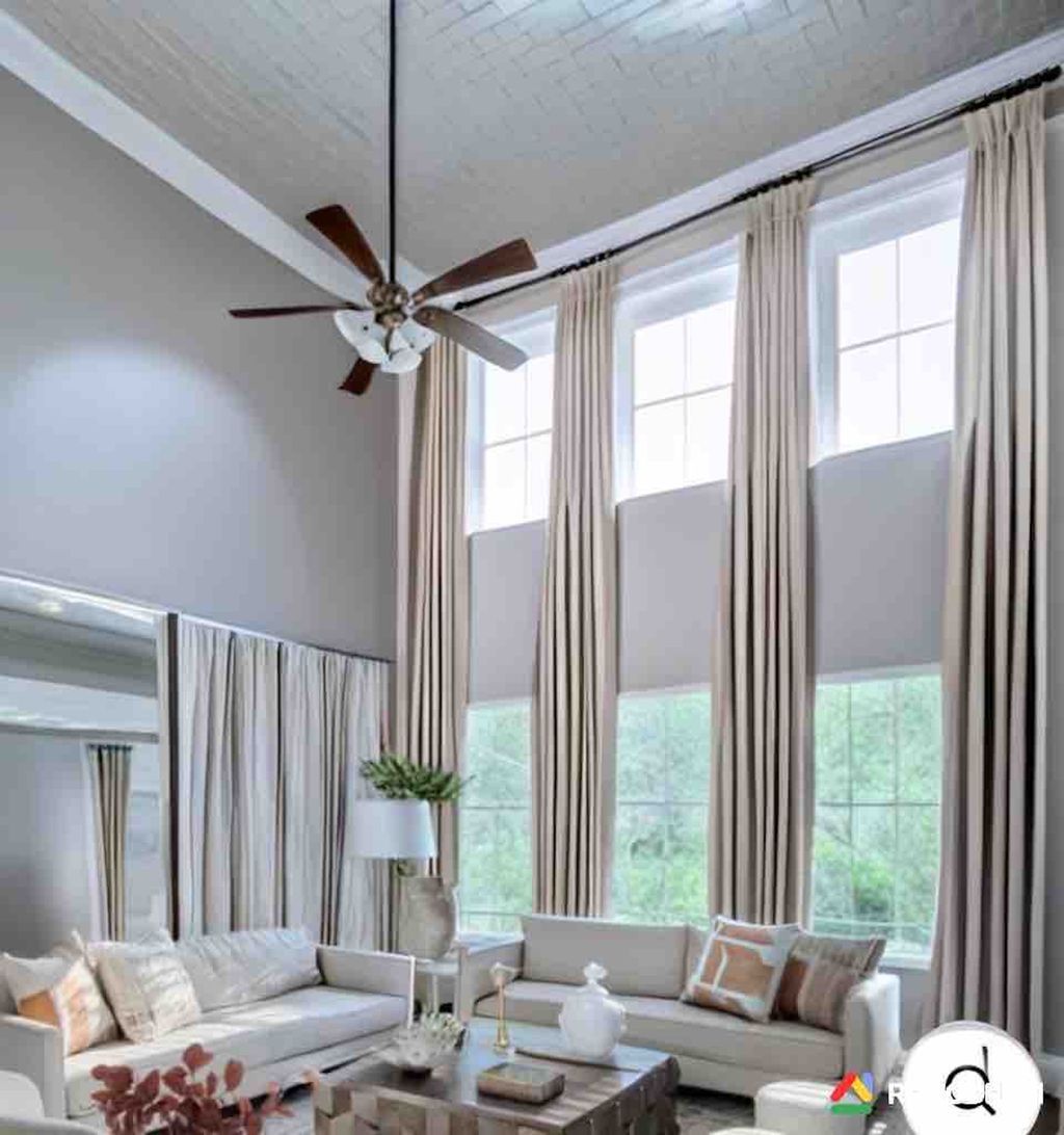 Bright living room with tall windows, white sofas, beige curtains, and a high ceiling with a ceiling fan