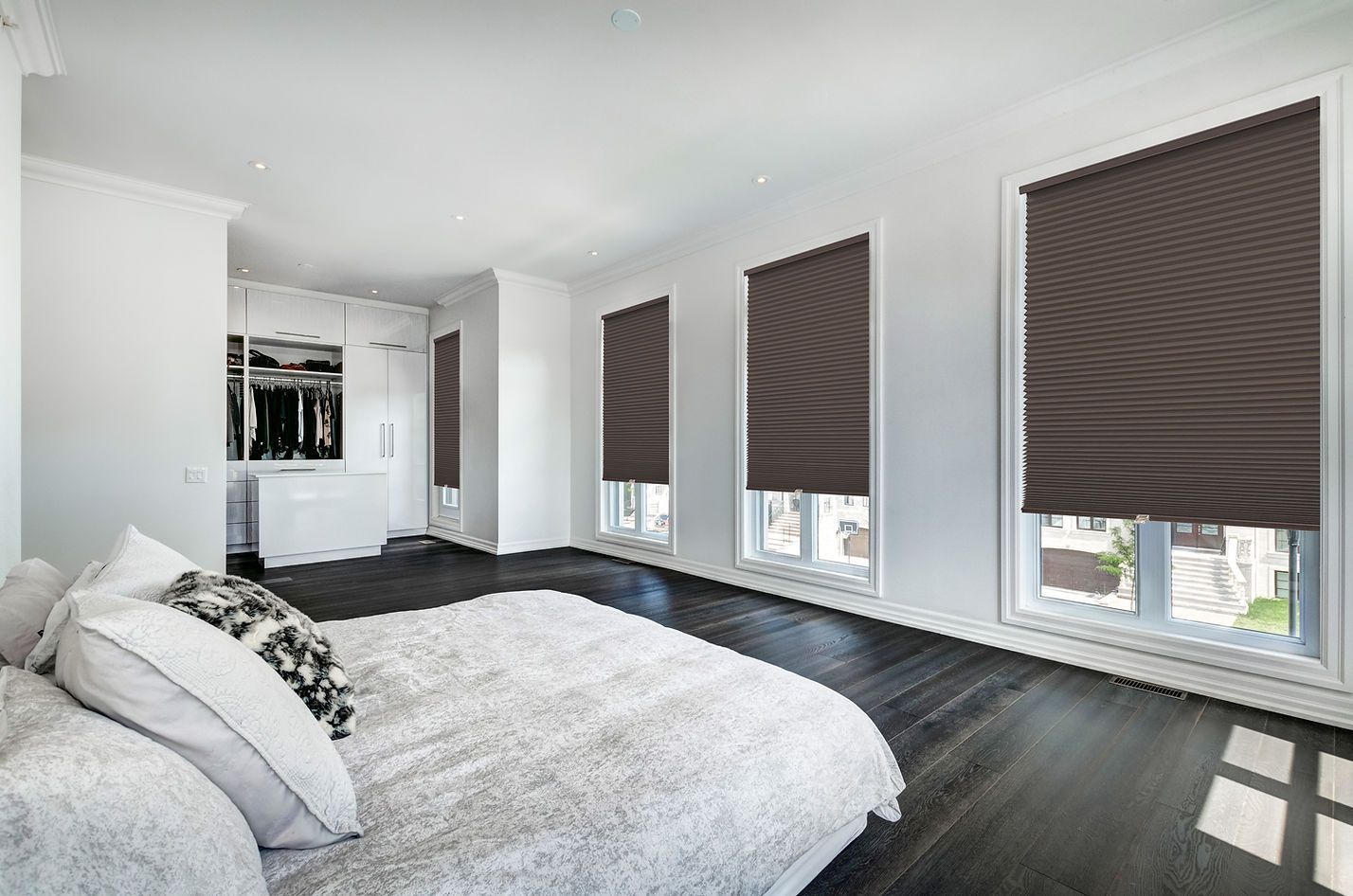 Bright bedroom with white bedding, dark floor, and large windows with closed blinds
