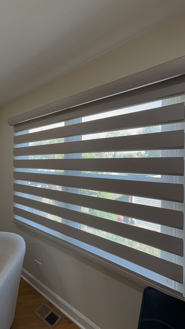 Closed zebra blinds filtering sunlight in a room near a window and white chair.