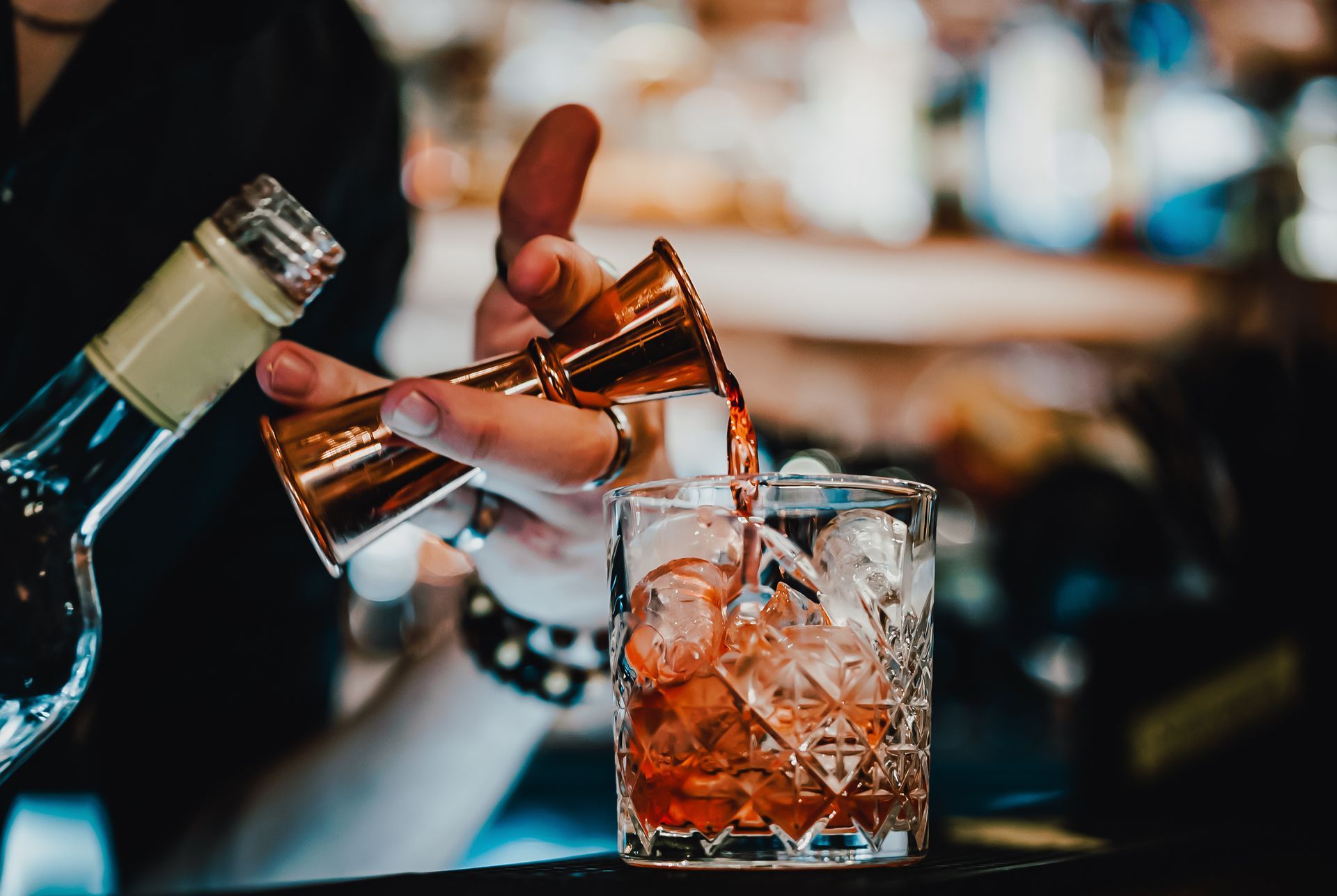 Two amber cocktails with rosemary sprigs and ice on a reflective bar, with blurred bottles and a lemon half behind them