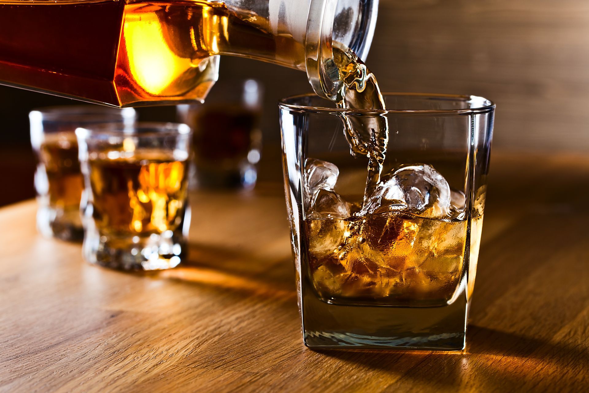 Whiskey being poured over ice into a tumbler on a wooden table with two glasses in the background