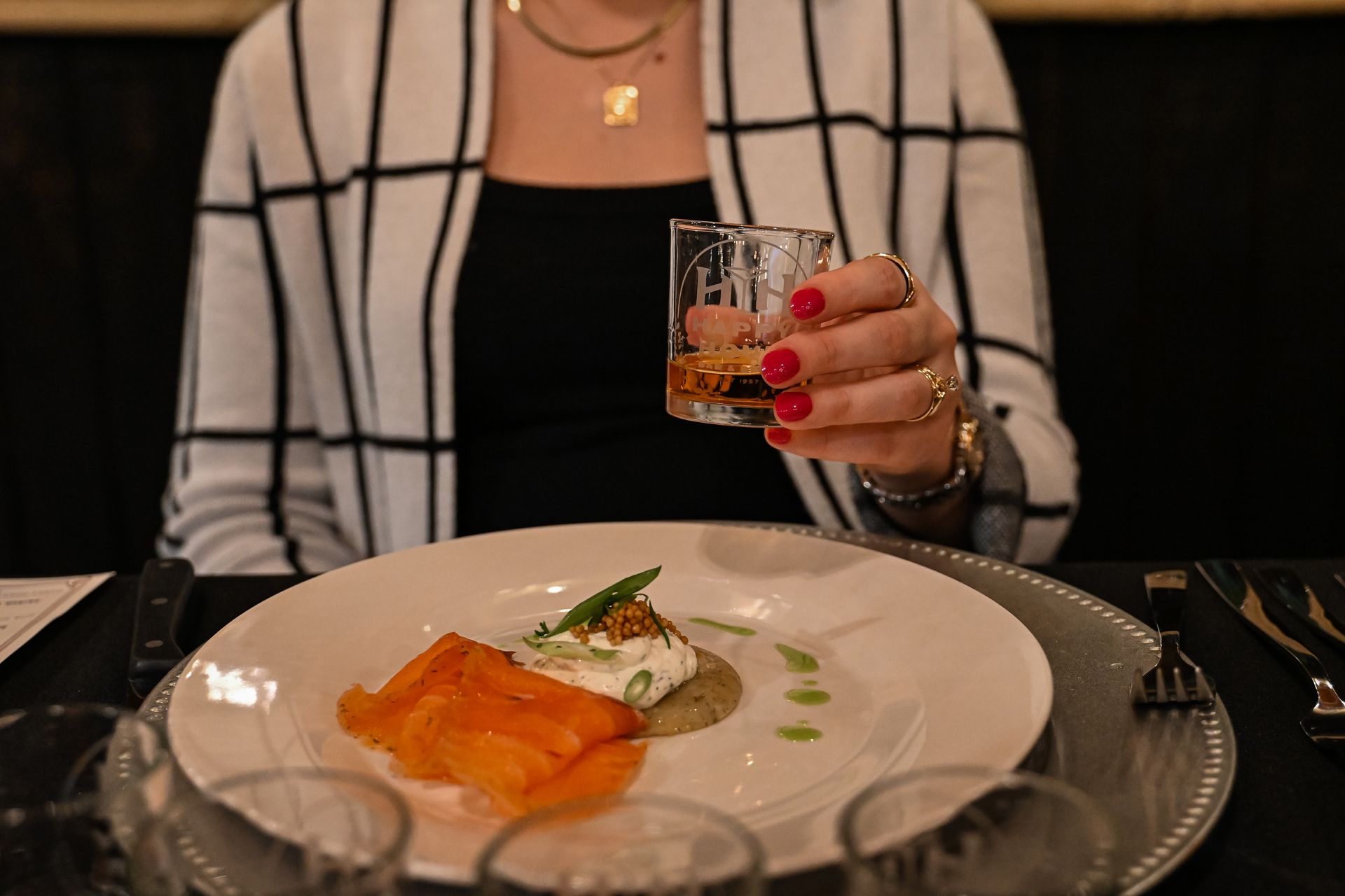 A woman is sitting at a table holding a glass of whiskey and a plate of food.