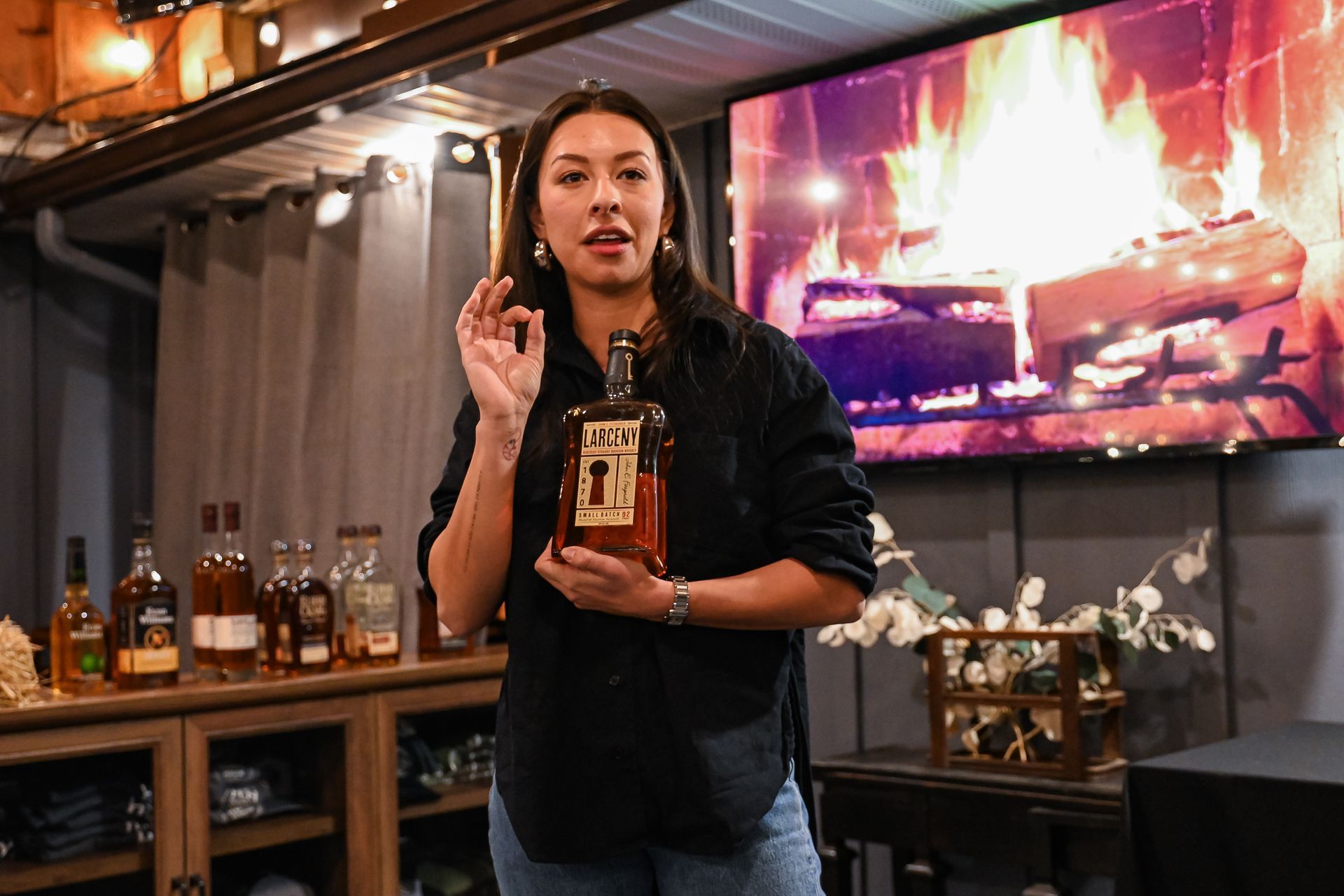 A woman is holding a bottle of whiskey in front of a fireplace.