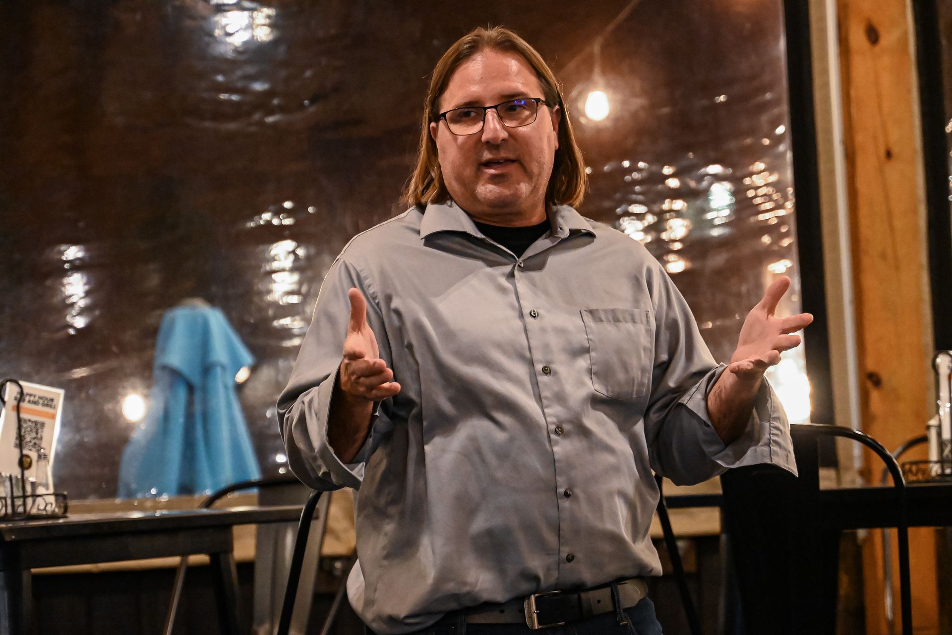 A man with long hair and glasses is standing in front of a table with his hands outstretched.