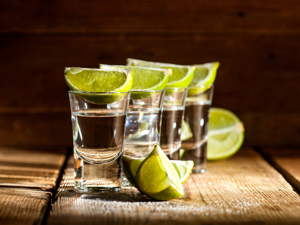 Tequila shots with lime wedges on a rustic wooden surface, illuminated by warm light.