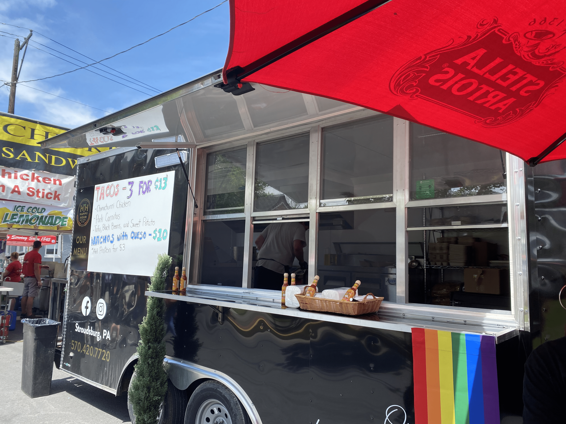 Happy Hour Bar & Grill food truck with a rainbow flag on the side is parked next to a red umbrella.