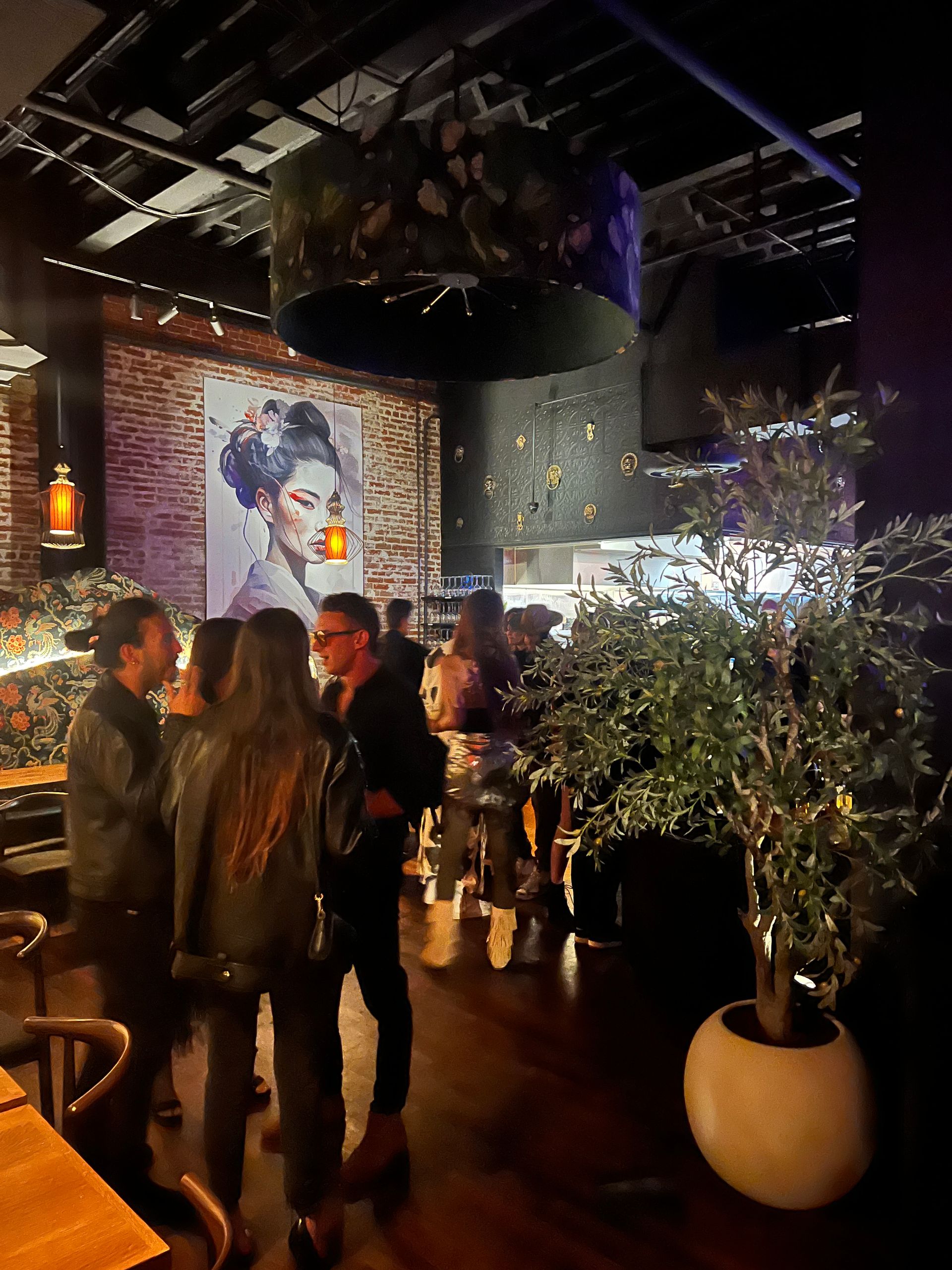 A group of people are standing in a restaurant with a painting of a geisha on the wall.