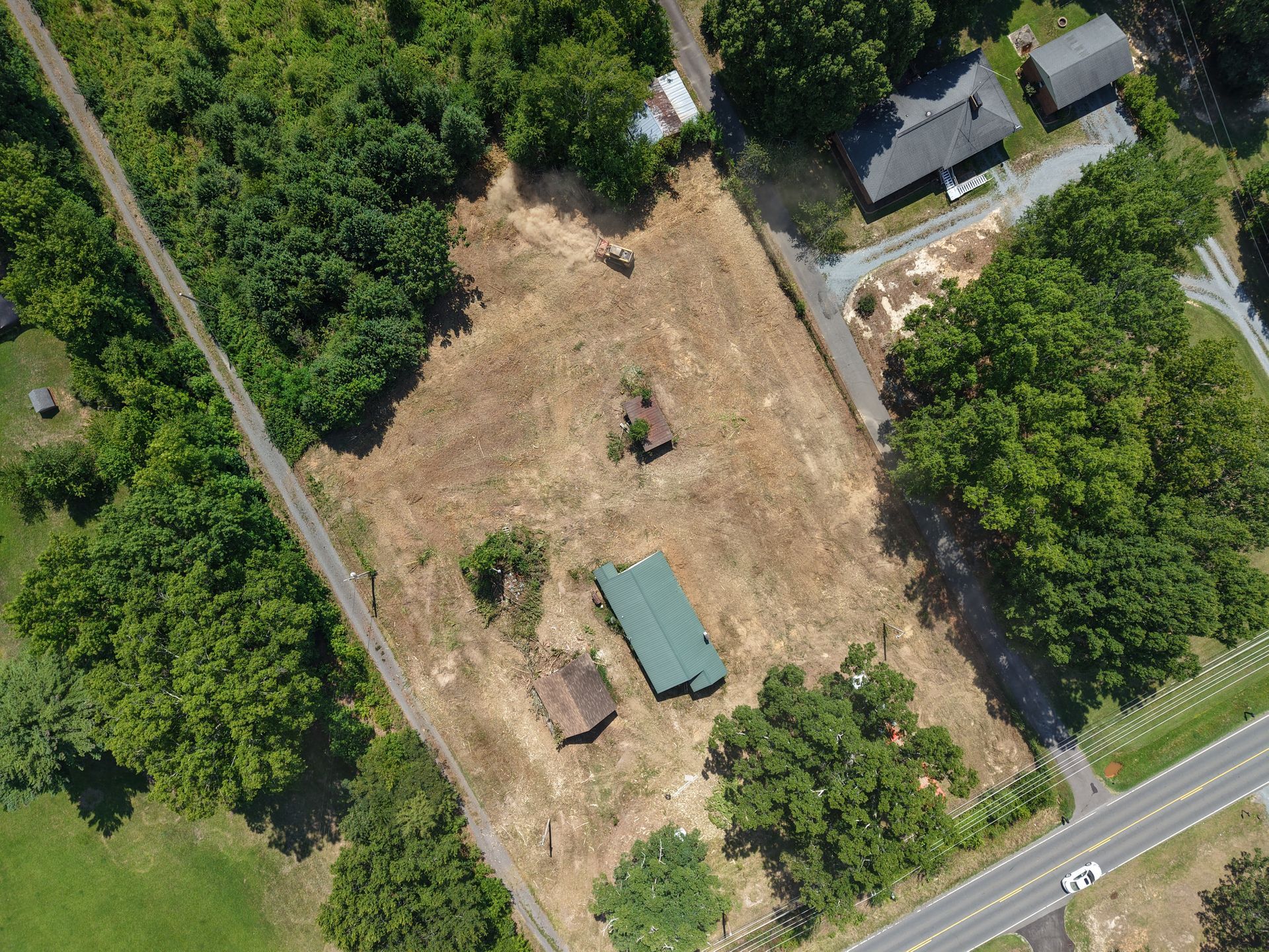 Aerial view of a rural property with a tan cleared field, small buildings, and surrounding green trees near a road