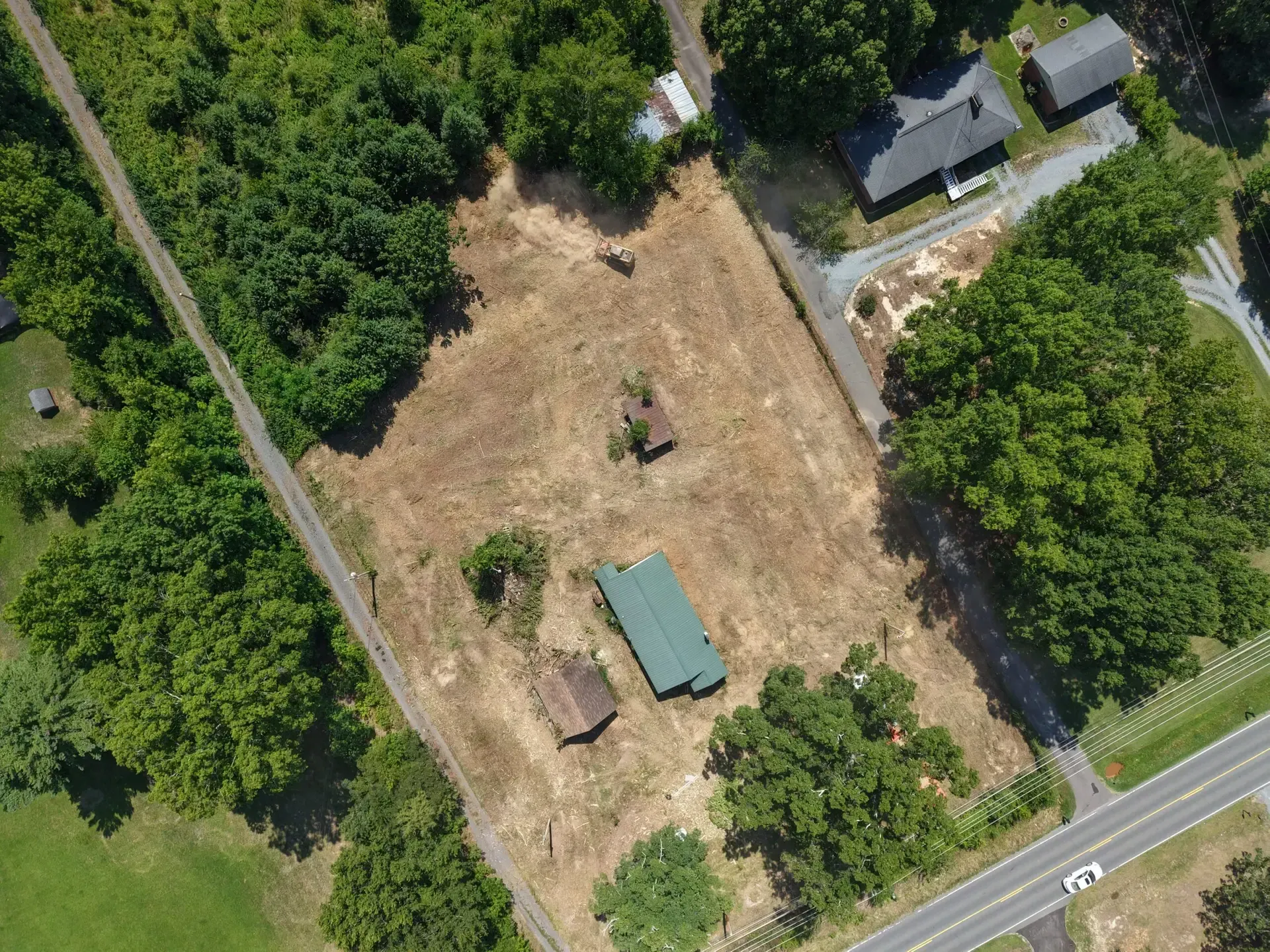Aerial view of a rural property with a house, shed, open dirt lot, and surrounding trees beside a road