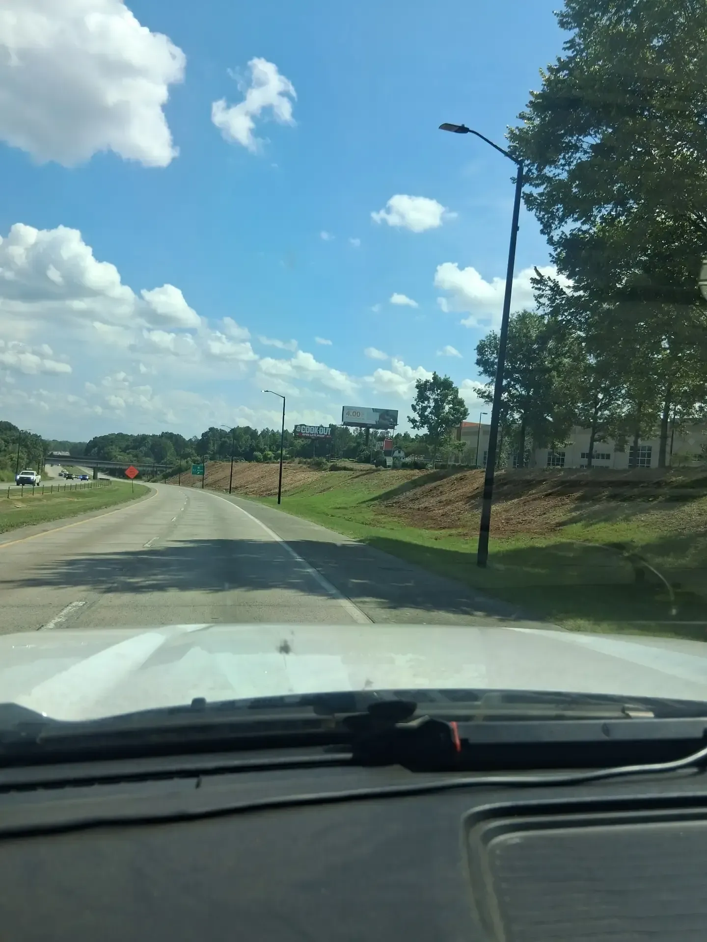 View from a car on a sunny road with trees, grass, and a cloudy blue sky ahead