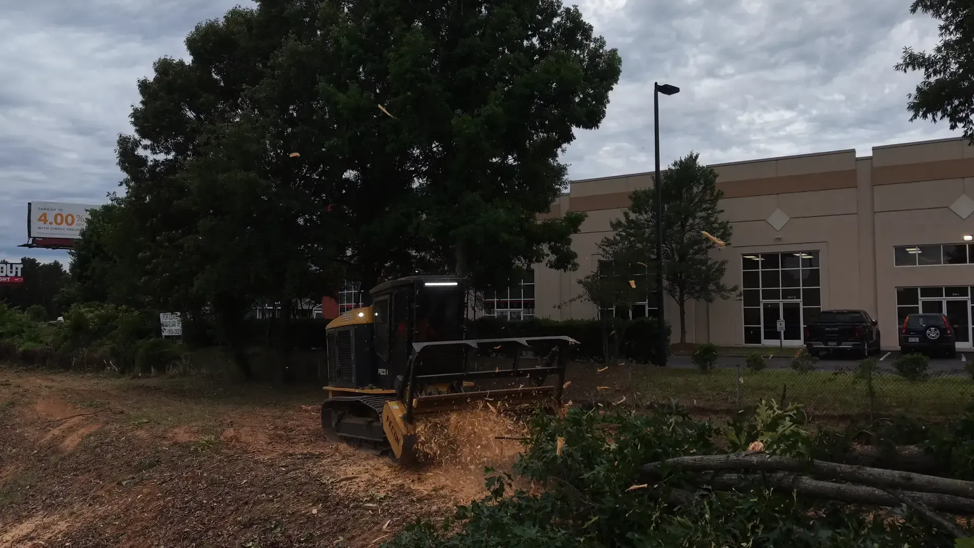 Construction equipment clearing mulch near a beige building under cloudy skies