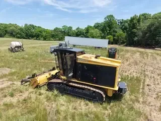 Yellow tracked tractor mower in a grassy field with trees and a white tank in the background
