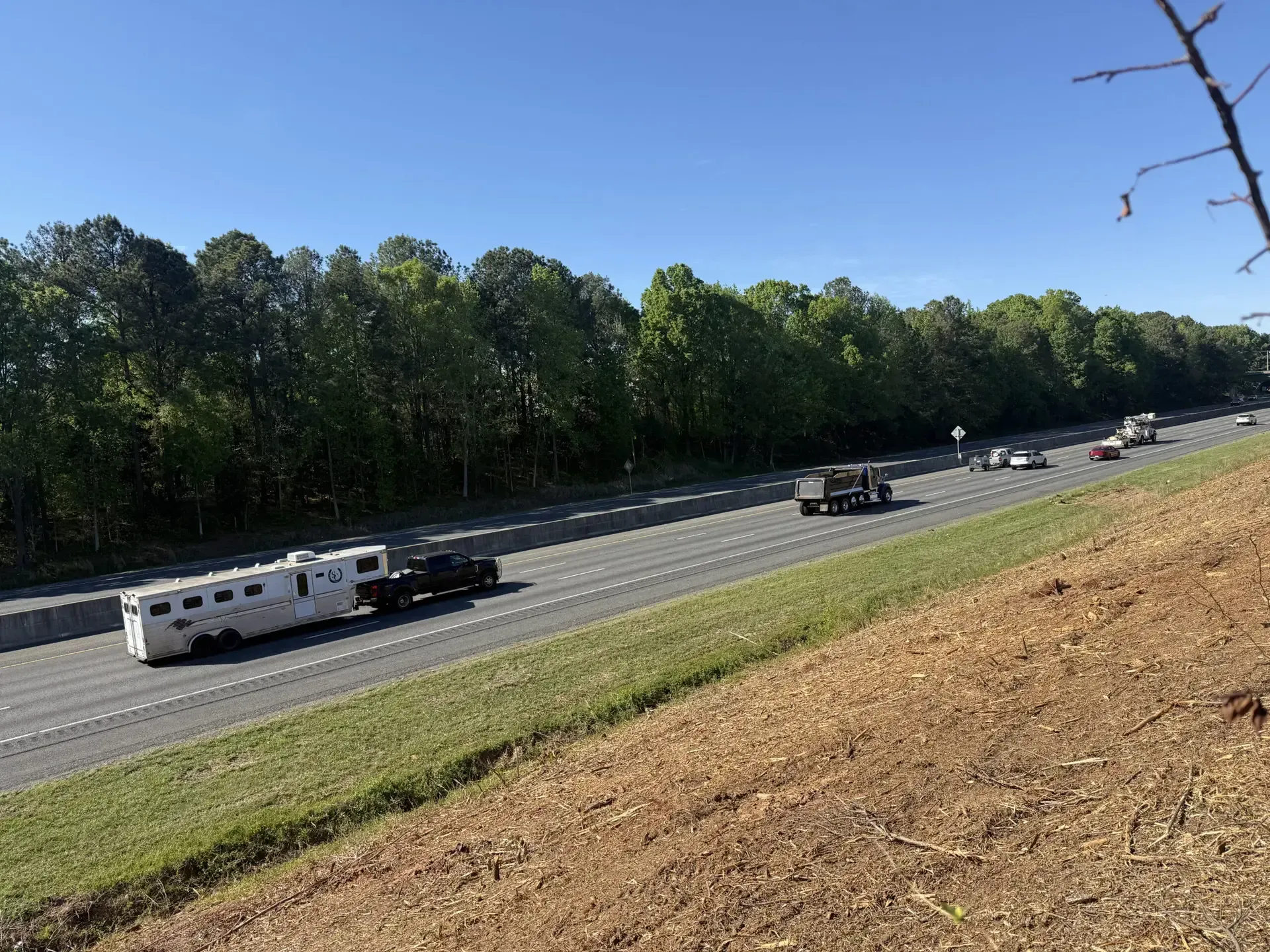 Cars and a white RV travel along a highway beside a wooded embankment under a clear blue sky