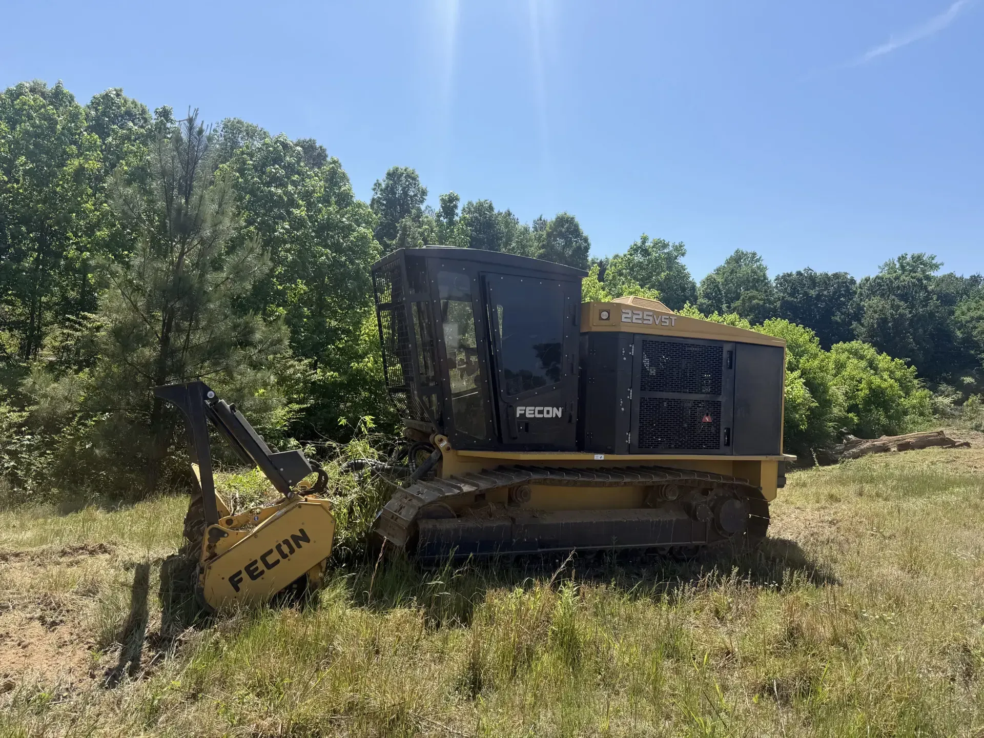 Yellow excavator in grassy field beside trees under a blue sky