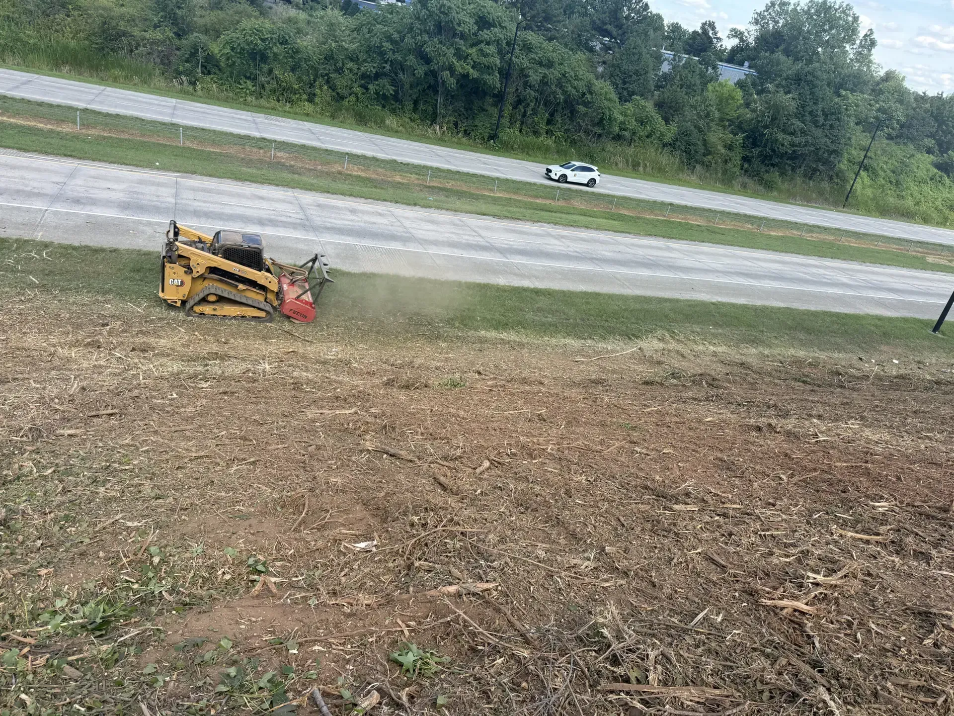Yellow skid steer mowing a grassy roadside slope near a highway with trees in the background