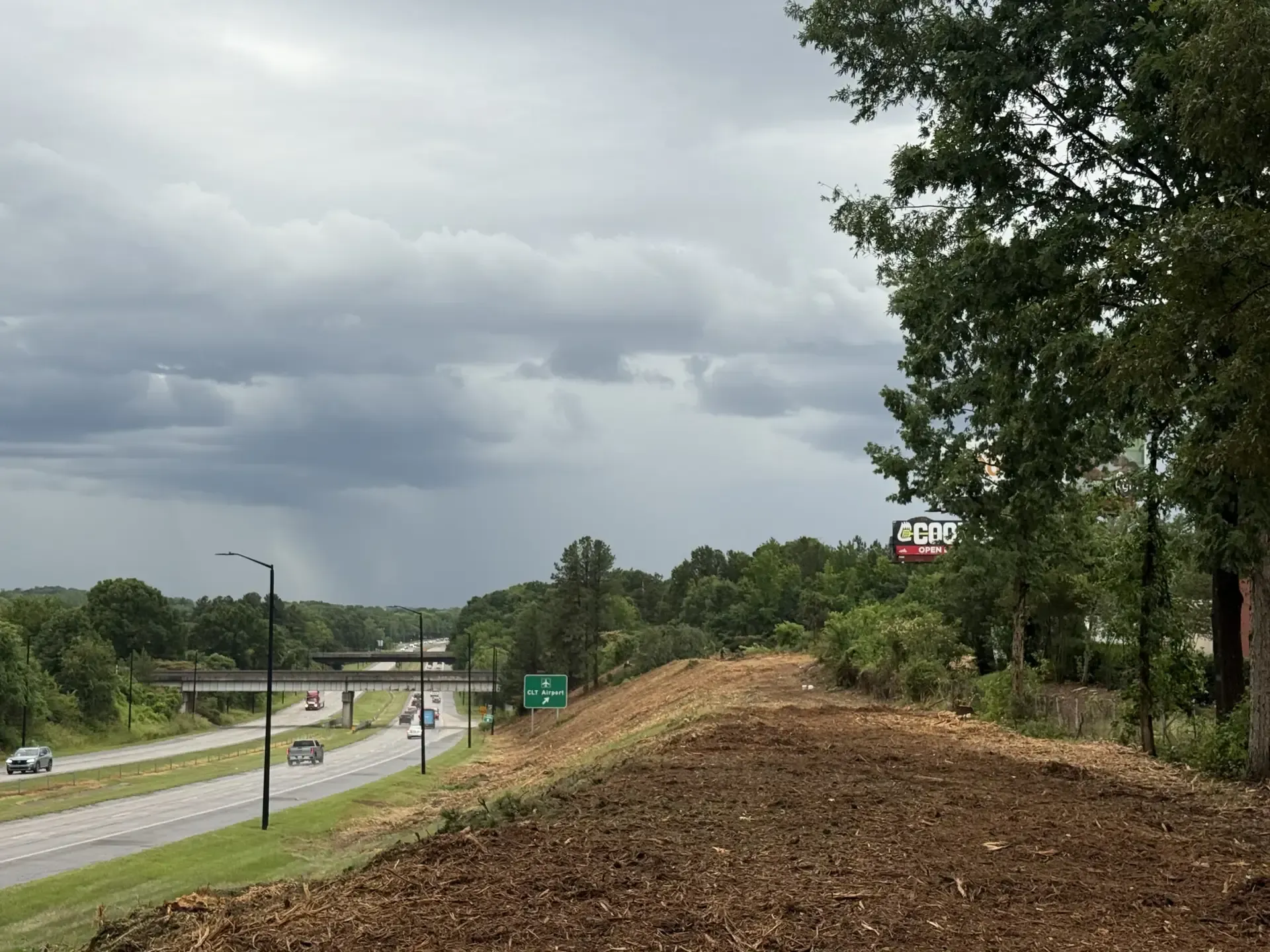 Highway beside a wooded hillside under cloudy skies, with a dirt embankment in the foreground