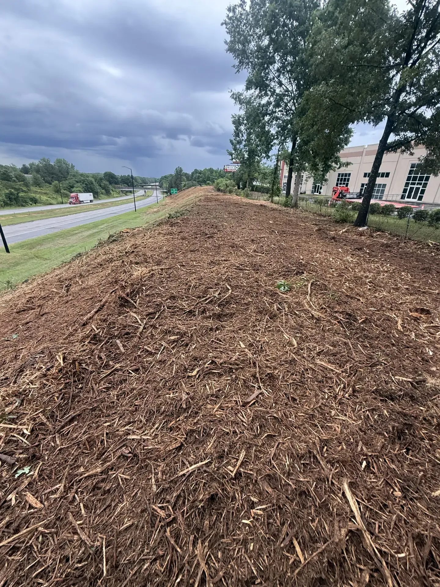Woodchip-covered embankment beside a road under cloudy skies, with trees and a building nearby