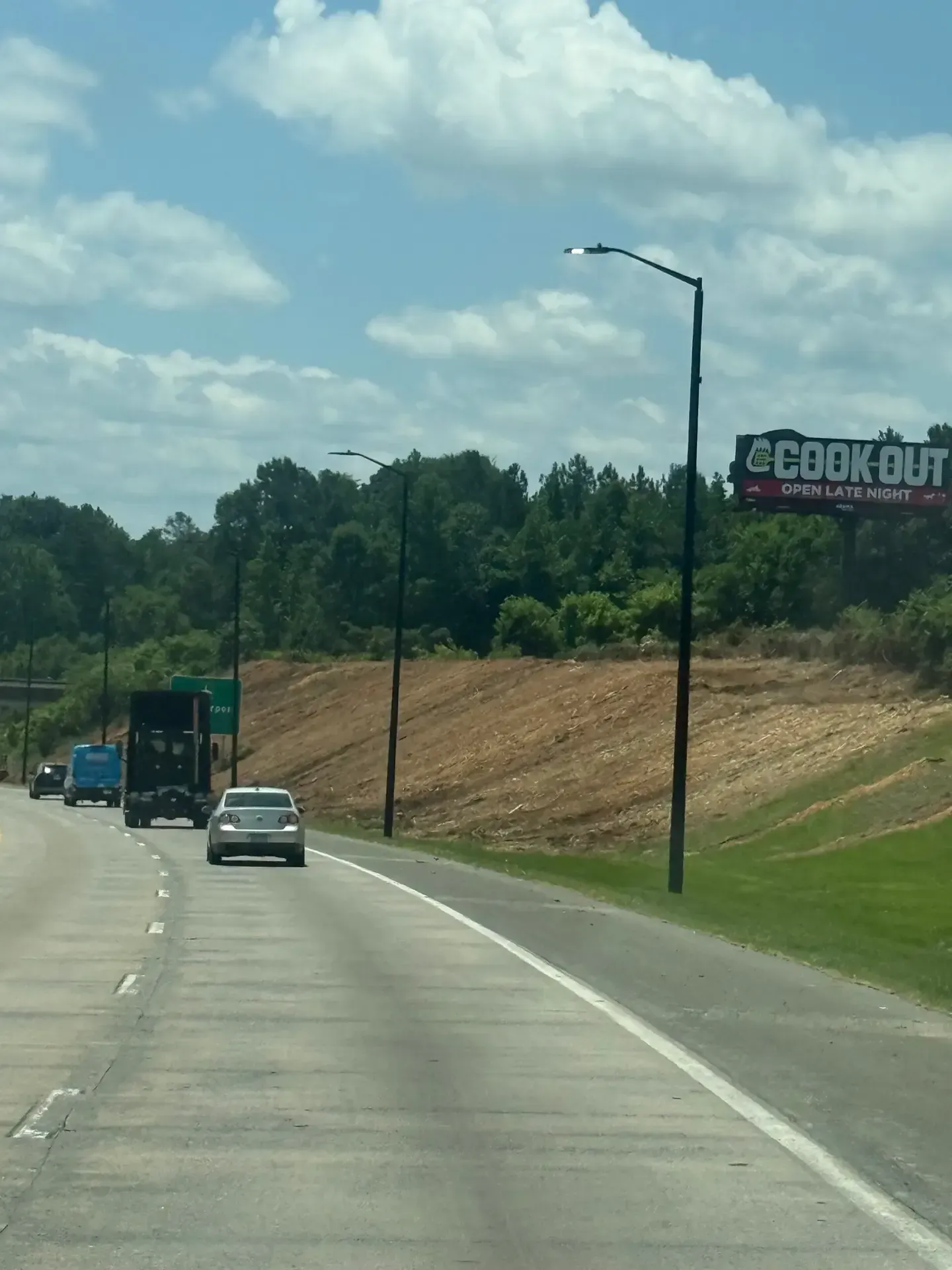 Highway with cars driving past a wooded embankment and a large “BROWNOUT” sign under a cloudy sky