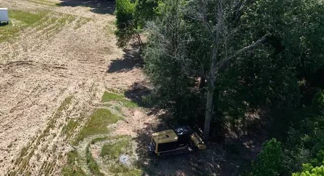 Aerial view of a dirt field beside trees, with a small yellow vehicle parked in the shade.