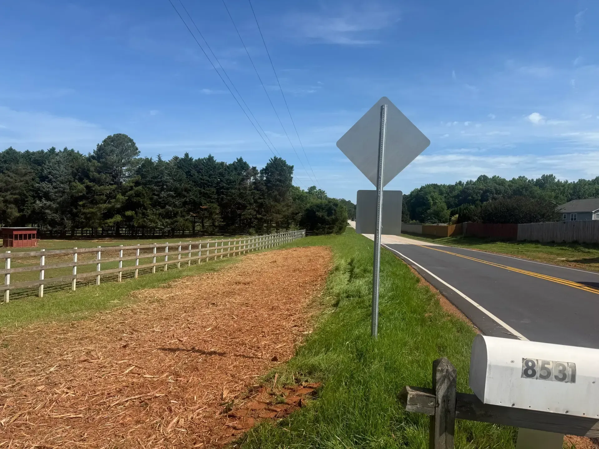 Rural road with a blank signpost, grassy shoulder, wooden fence, and trees under a blue sky