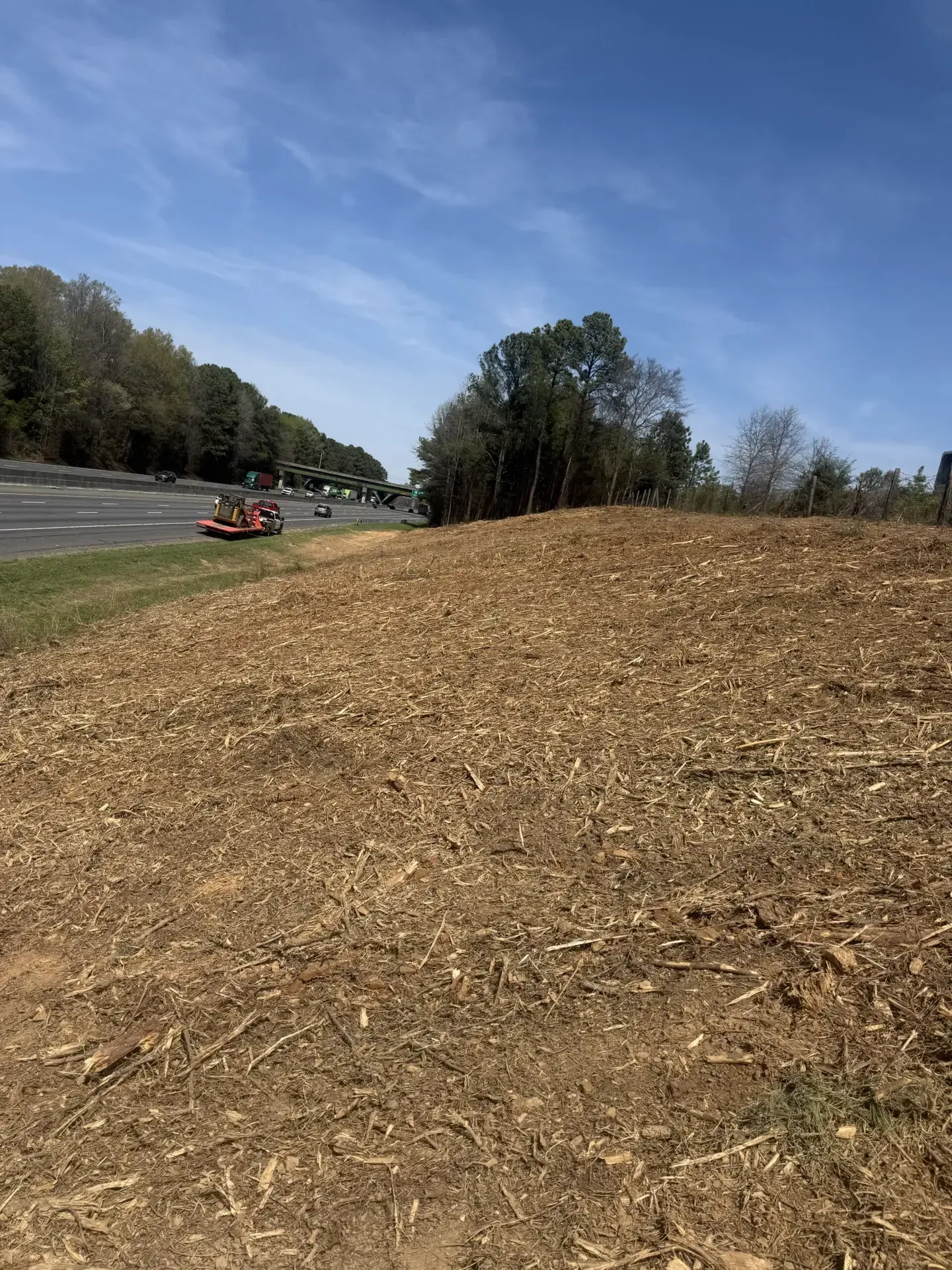 Dry field with a red tractor in the distance under a blue sky and trees along the edge