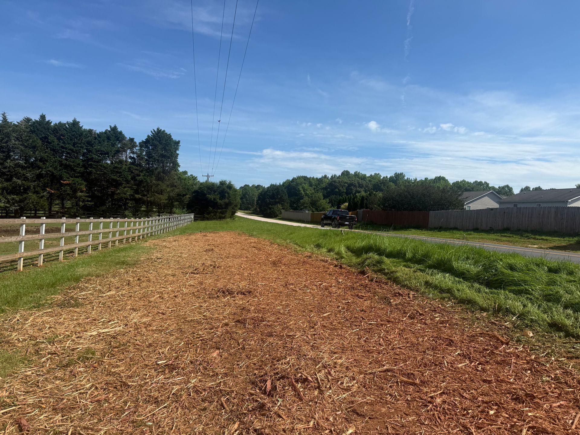 Woodchip path through a green field under a blue sky, with a fence and trees along the left side.