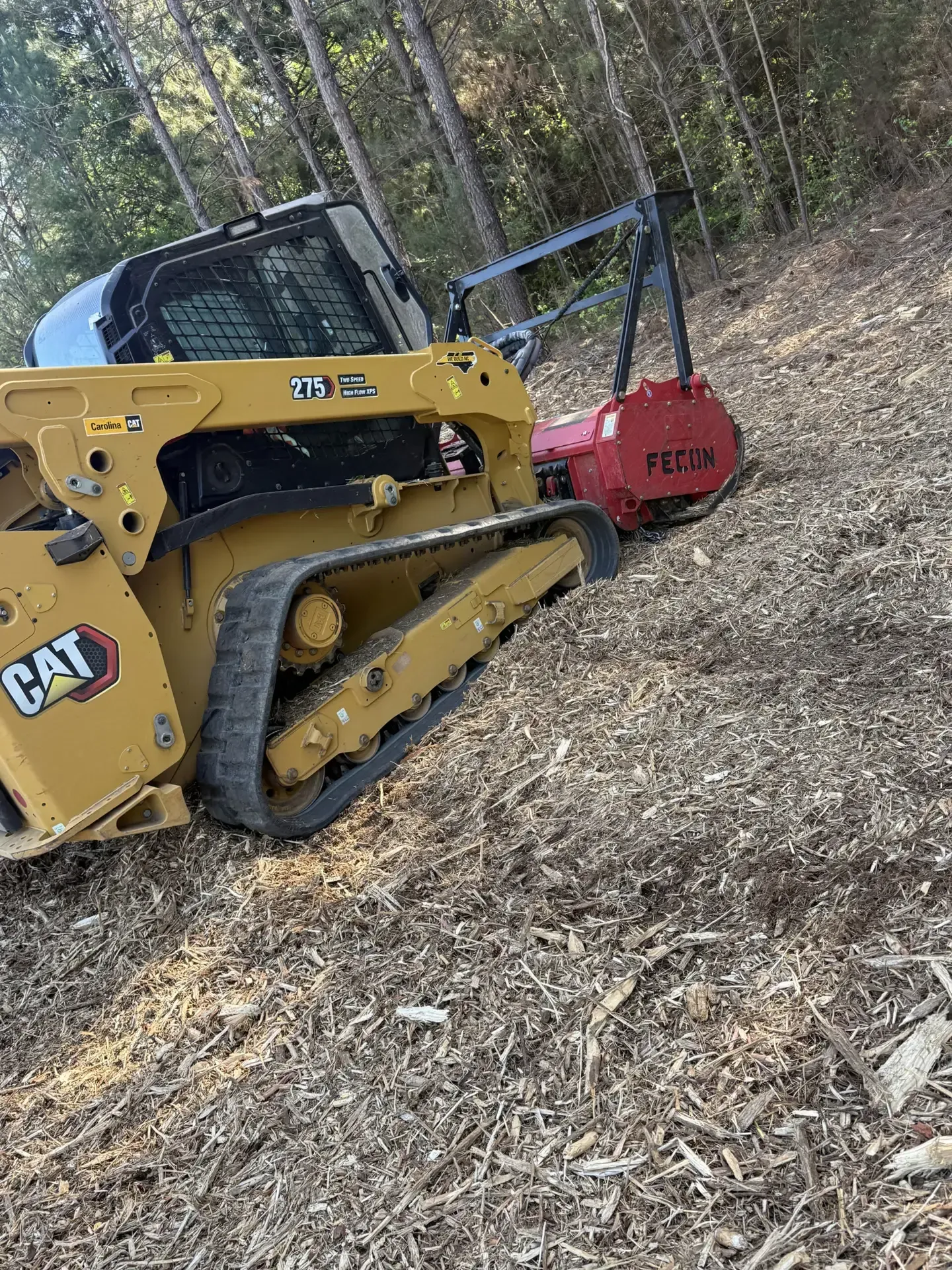 Yellow CAT skid steer pushing a red brush cutter through a wooded, leaf-covered slope