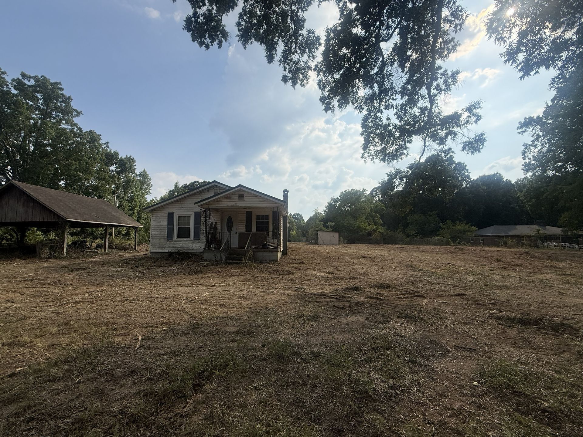 Wide dirt yard with a small house, shed, and trees under a cloudy sky.