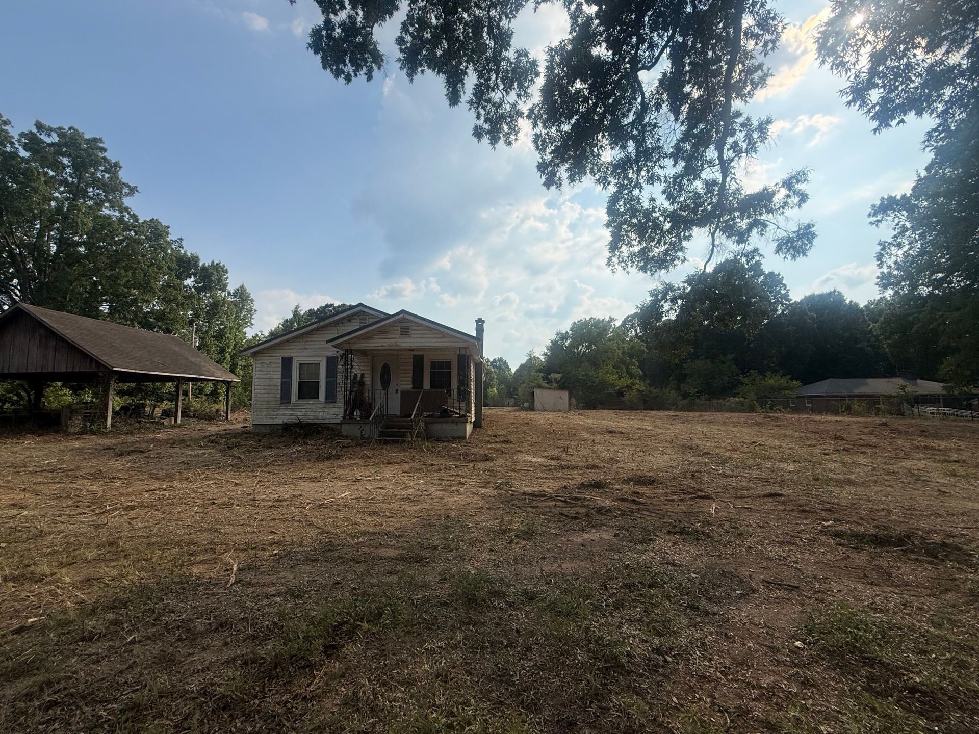 Small white house in a dry, open yard with trees and a gazebo under a bright sky