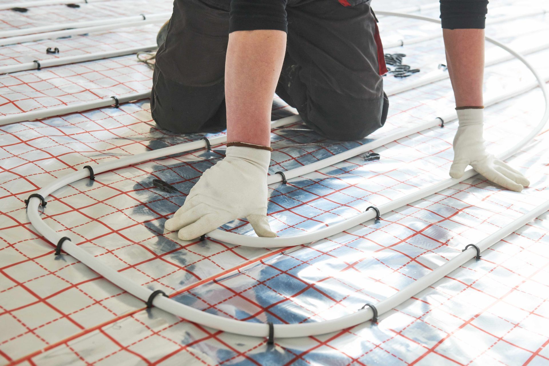 Person installing radiant floor heating system, using white tubing and fasteners on a checkered surface. Person installing radiant floor heating system, using white tubing and fasteners on a checkered surface.